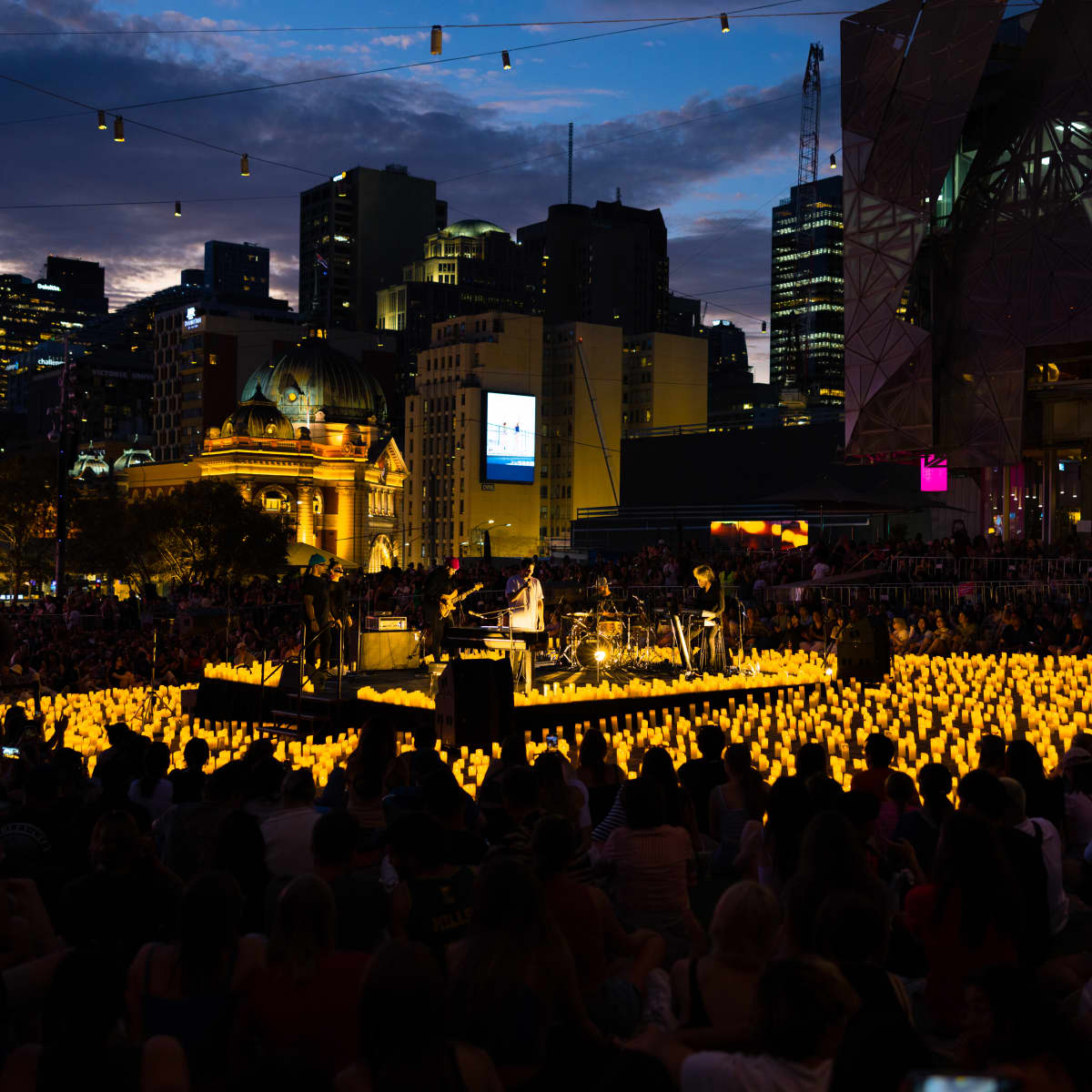 photo at dusk of a candlelight concert at melbourne's Fed square, showing candles, silhouettes of a band and audience with the city skyline in the background