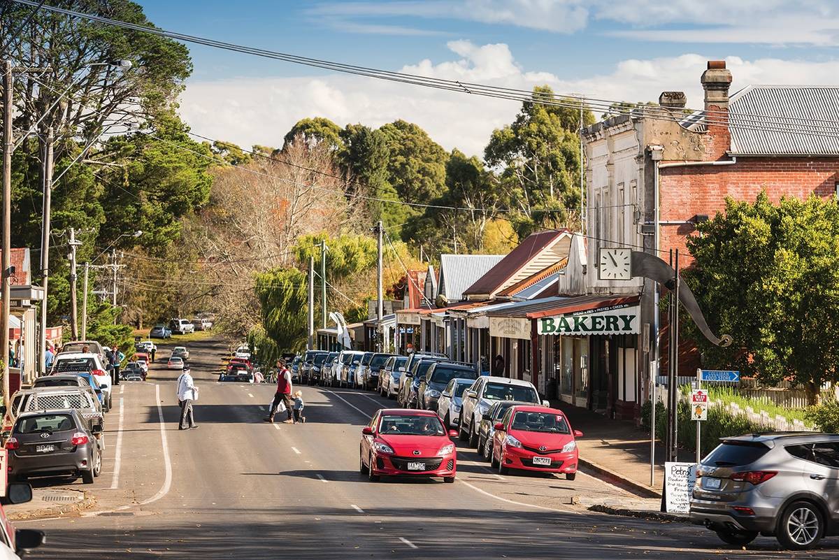 The main street of Trentham, featuring a line of parked cars and charming storefronts, including a bakery, set against a backdrop of leafy trees and a clear sky