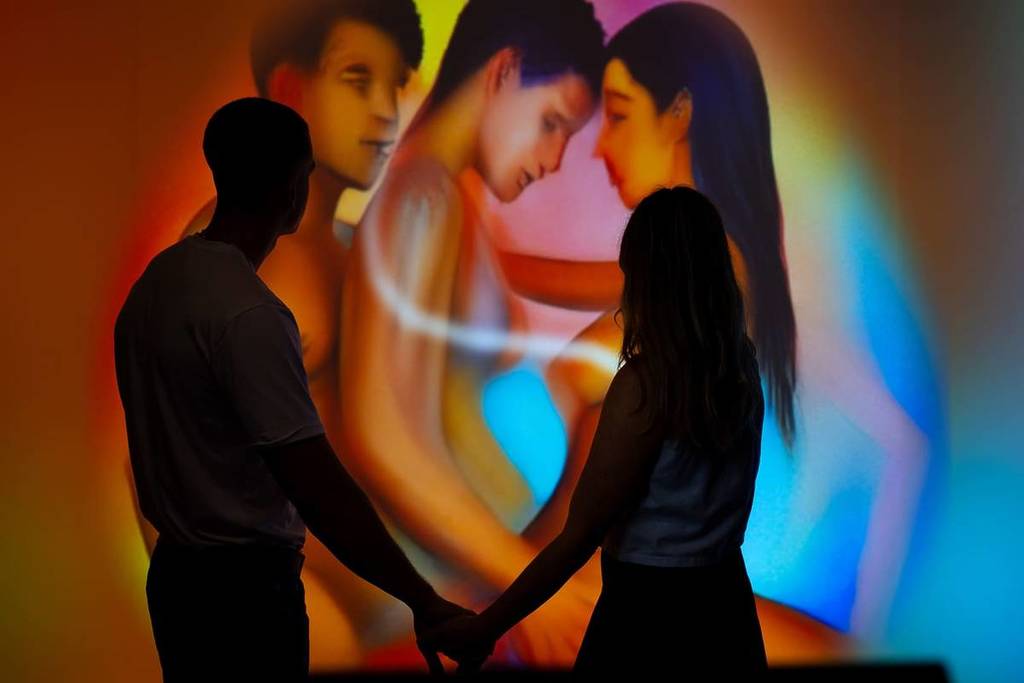The silhouettes of a man and a woman are seen holding hands in front of a piece of digital art depicting a threesome at the Museum Of Desire.
