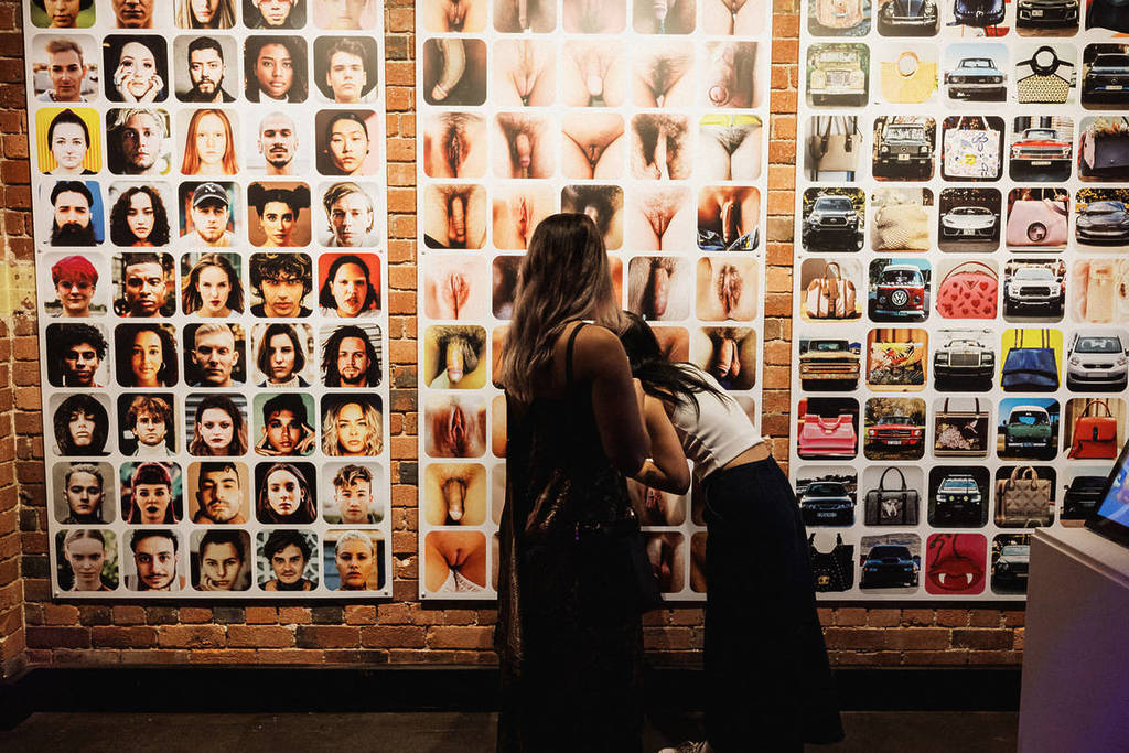 Two women stand in front of a wall with three panels of photos depicting people, genitalia and vehicles at the Museum Of Desire.