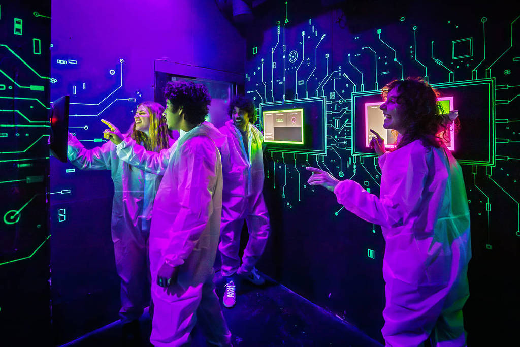 Four young adults in white jumpsuits stand in a room playing games on screens mounted on the wall at 123Boom!