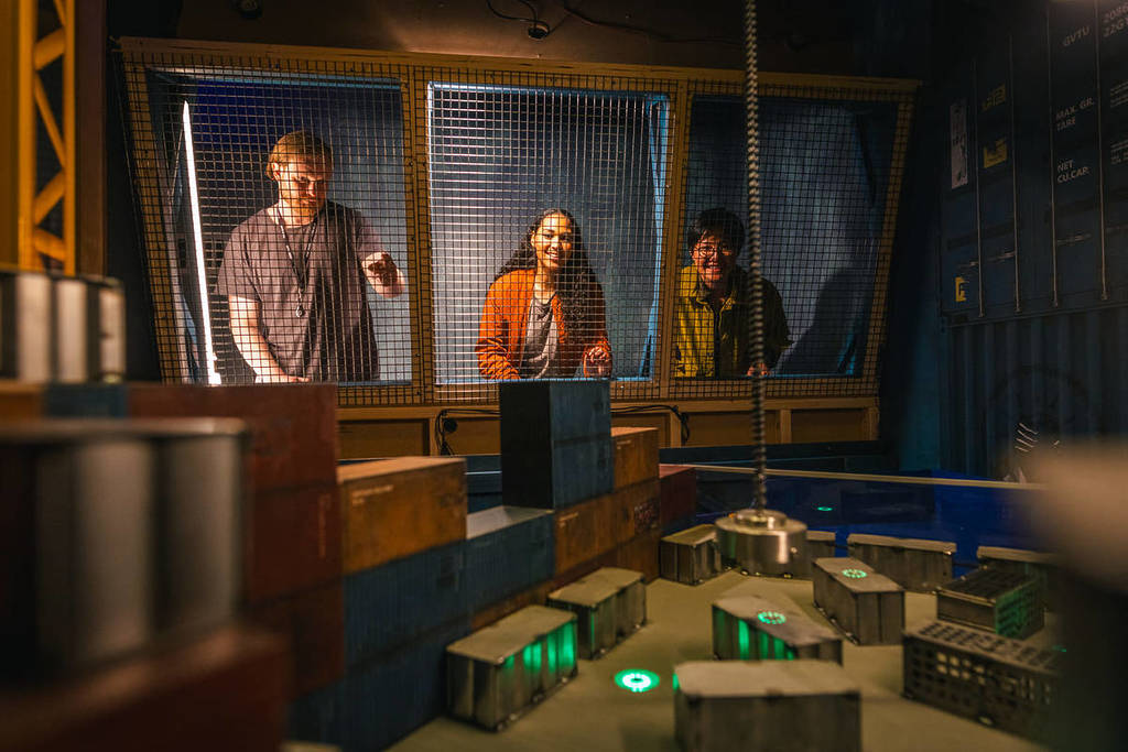 A young man and two young women stand behind a set of windows in one of the game cells at Prison Island. They are looking through the windows and pointing at the game inside.