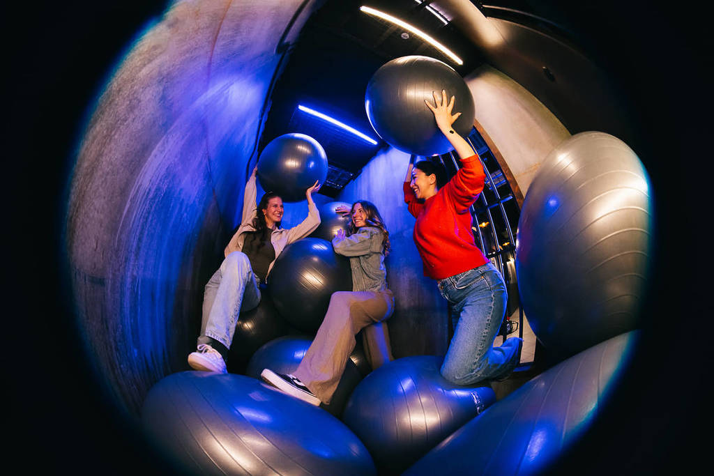 Three young women are in a game cell at Prison Island. They are standing on and holding up giant bouncing balls.