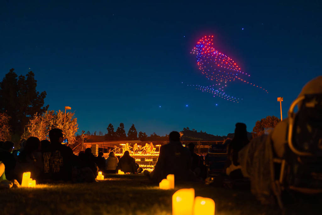 An orchestra surrounded by candles plays underneath the night sky, which is lit up with the image of a bird created by drones for DroneArt Show.