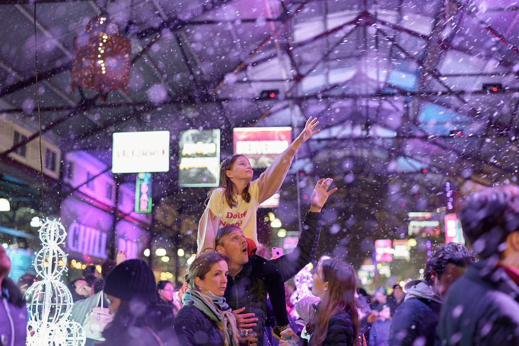 child in parent's shoulders reaching out to touch falling snow at Winter Night Market