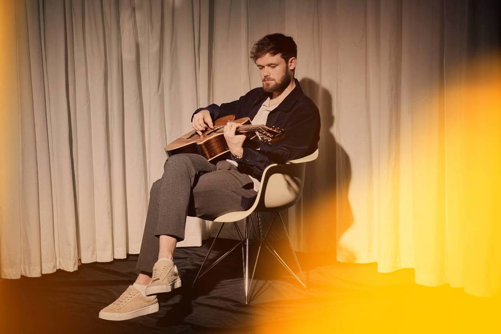 James TW sat on a chair and playing a guitar for the promotional Candlelight concert original session