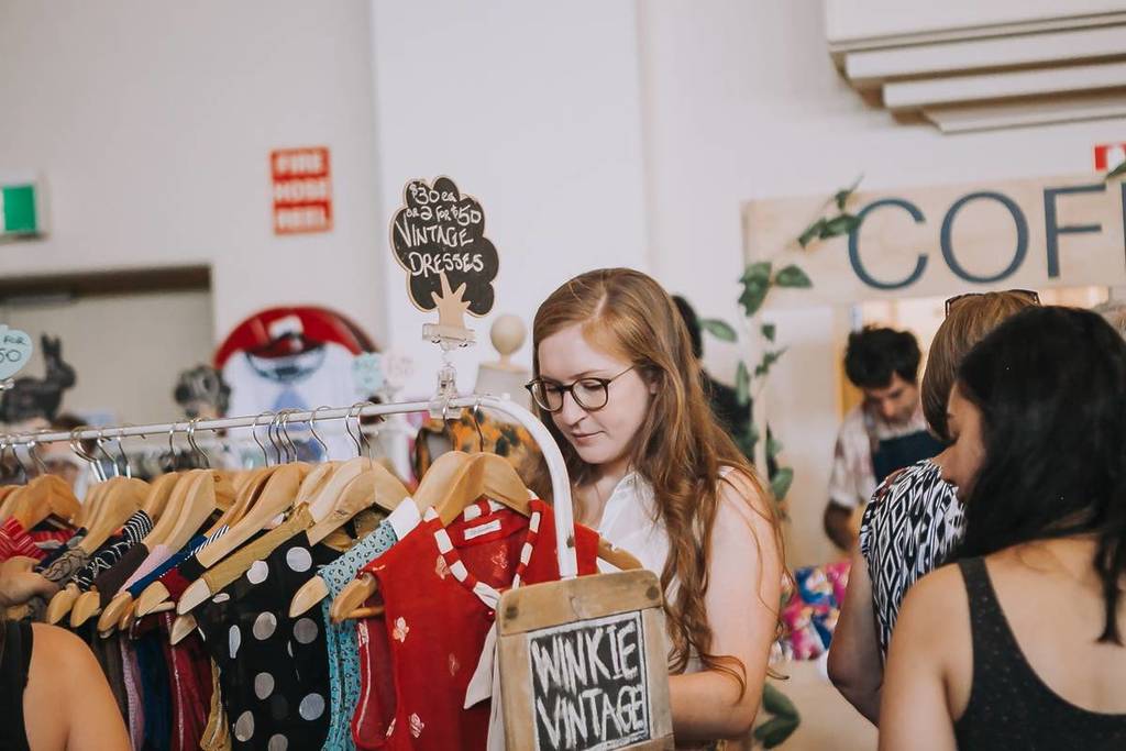 woman in glasses looking through a rack of vintage dresses at Round She Goes, with a coffee stall in the background