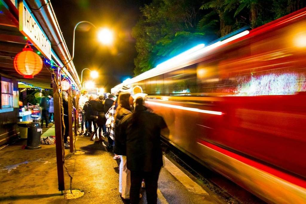 people standing on a platform at night while a train with lights rushes past, to depict the Walhalla Winter Light train experience