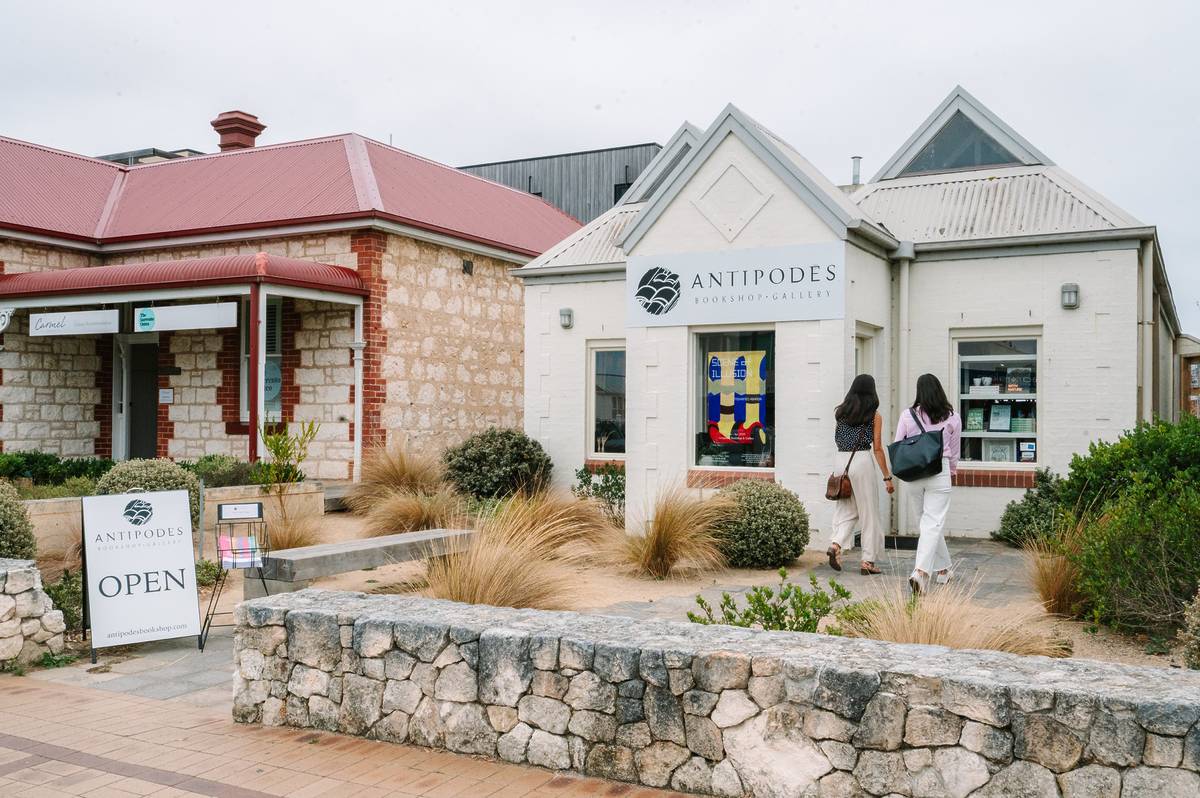 Two women walk toward Antipodes Bookshop & Gallery in Sorrento, Victoria, housed in a charming coastal-style building with a red tin roof and stonework facade