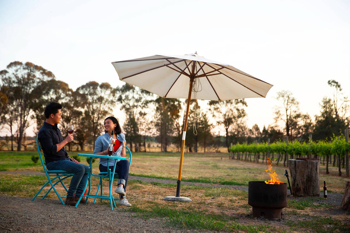 A couple enjoying wine at an outdoor table under a large white umbrella, with a fire pit nearby and vineyard rows in the background at sunset in Rutherglen, Victoria