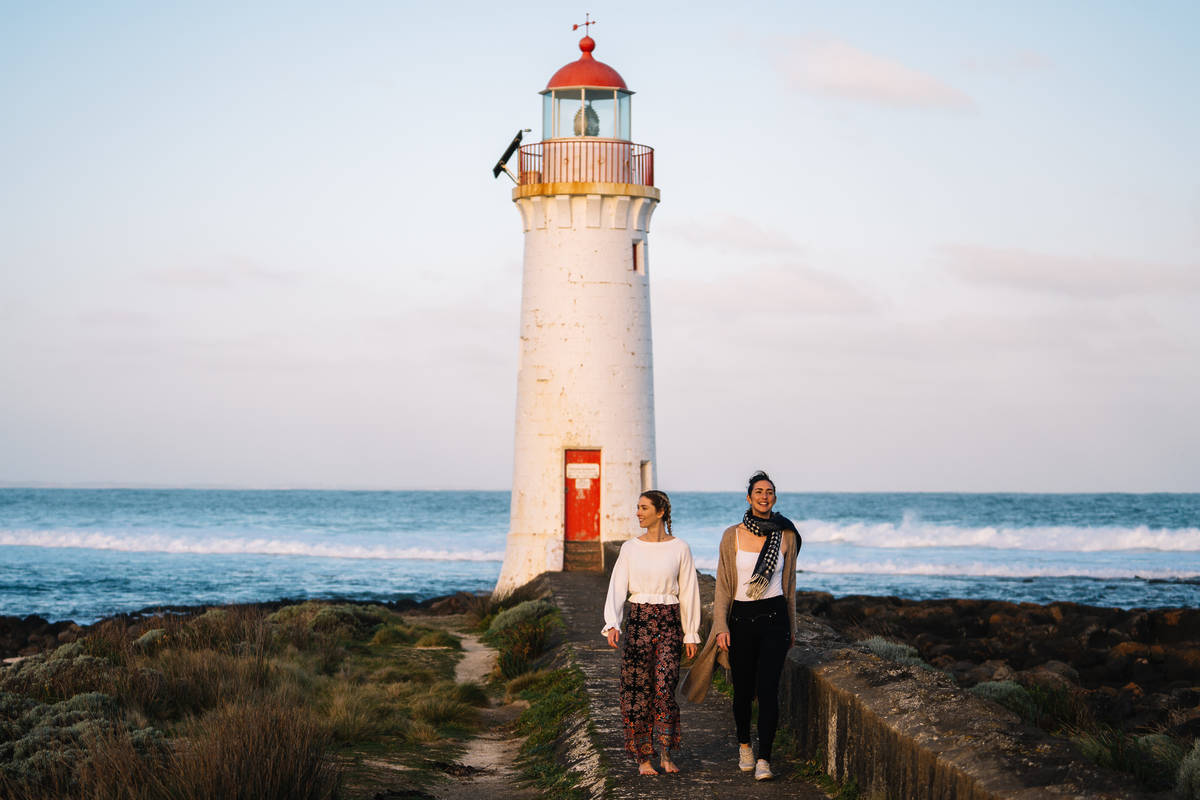 Two women walking along the trail to the Griffiths Island Lighthouse in Port Fairy, Victoria, with waves breaking in the background