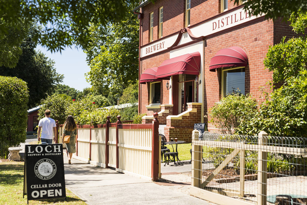 A couple walks toward the red-brick Loch Brewery & Distillery, with red awnings and a "Cellar Door Open" sign out front