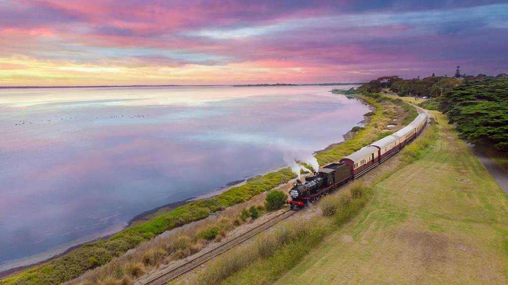 aerial view of The Blues Train moving along the coast at sunset, with pretty pink and blue colours on the still ocean water