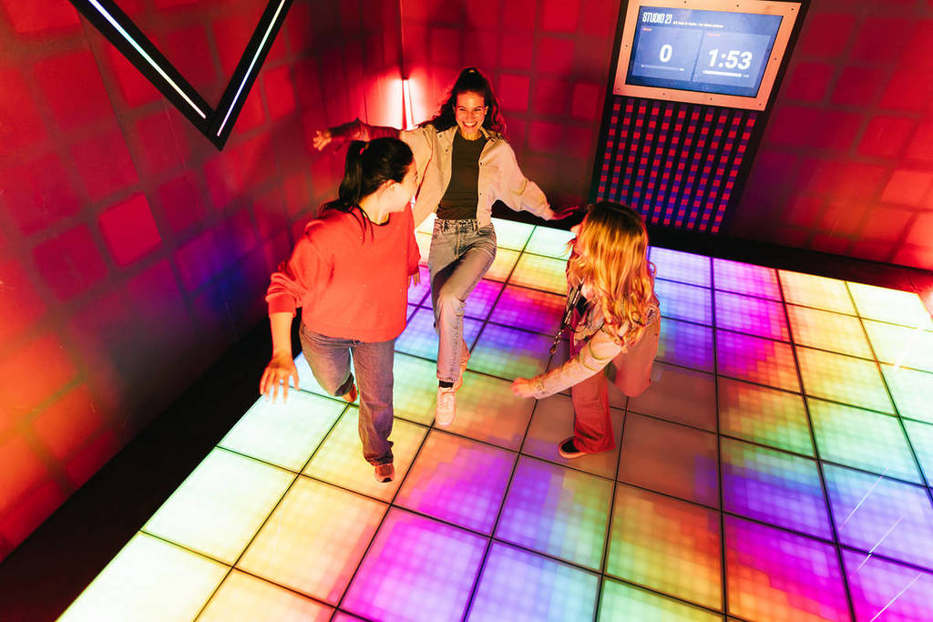 Three girlfriends have fun on a tiled floor that is lit up with colours at Prison Island.