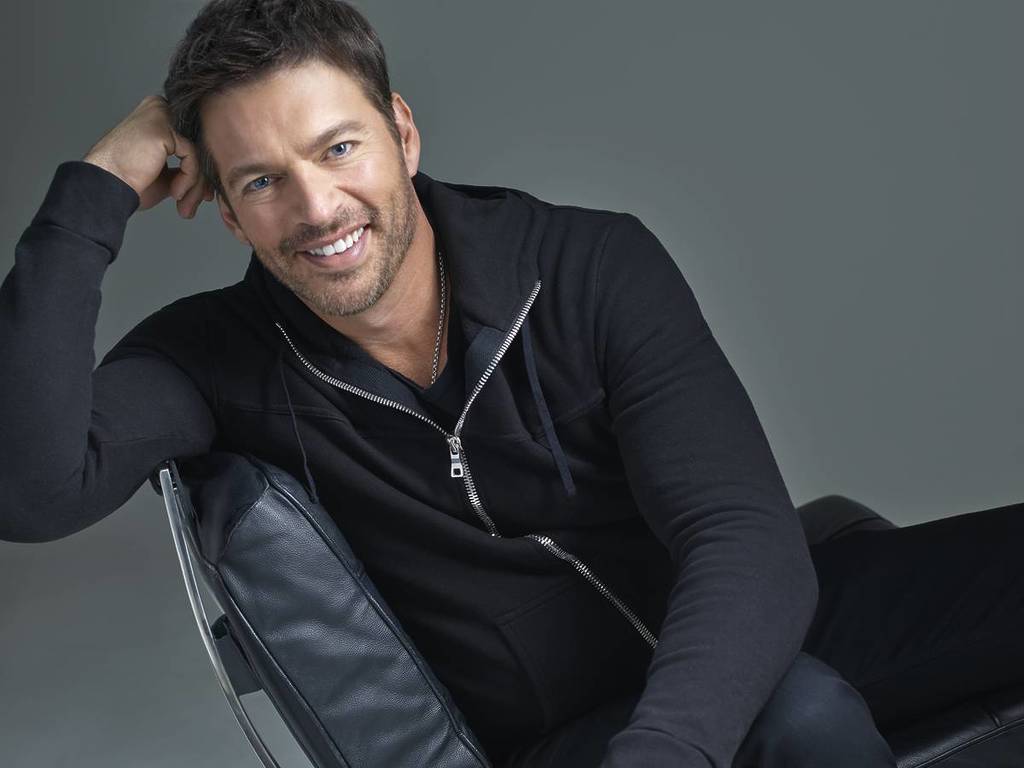 Harry Connick Jr in a zip up hoodie on a chair smiling at the camera