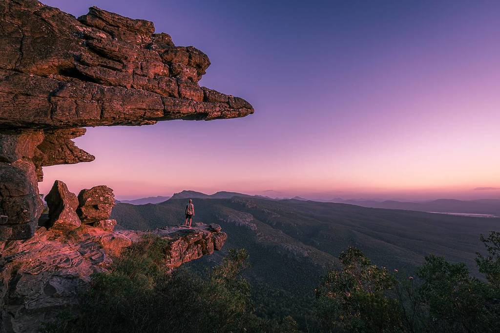 Person standing on a dramatic rock ledge at sunset, overlooking the vast mountain ranges of the Grampians National Park in Victoria
