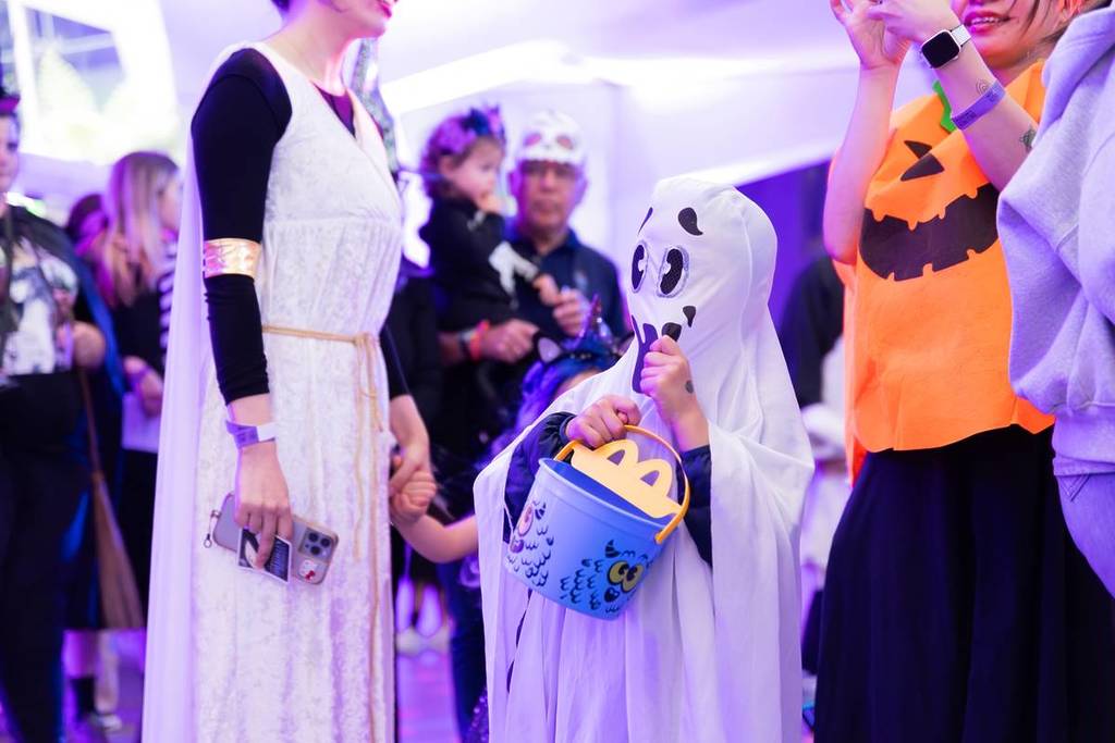 child dressed as ghost carrying a trick or treat bucket surrounded by adults in costume for Halloween at the Museum