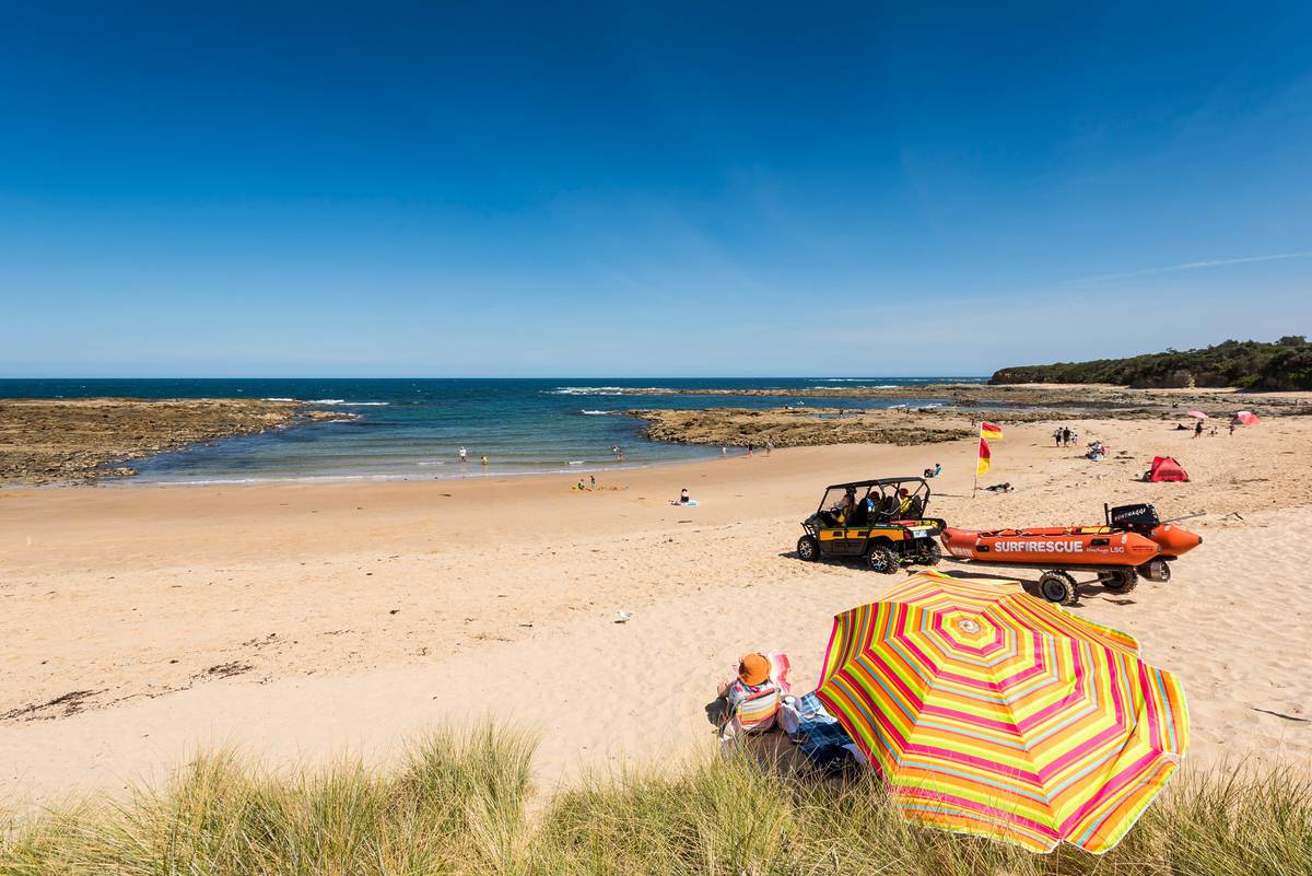 Cape Paterson Bay Beach, with golden sand, clear blue water, and a colourful beach umbrella in the foreground beside a surf rescue vehicle