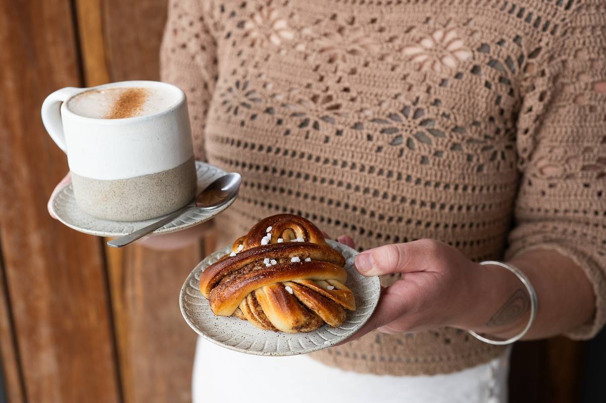 A person holding a coffee and a cinnamon bun served on ceramic plates at Zeal & Flow cafe in Cape Paterson