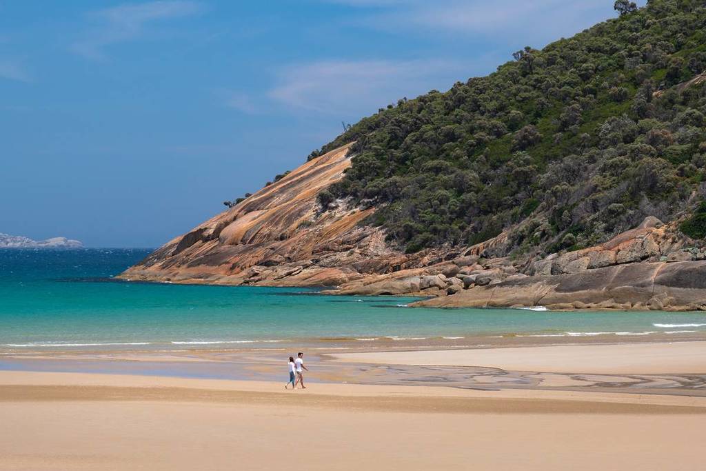A couple walking along the beach at Wilsons Promontory, with dramatic rocky cliffs and clear turquoise waters in the background