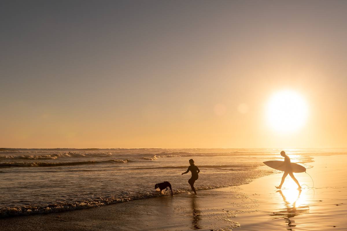 Silhouette of a person walking along the beach with a dog at sunset, and a surfer carrying their board along the shore in Venus Bay, one of the most underrated cheap summer getaways in Victoria