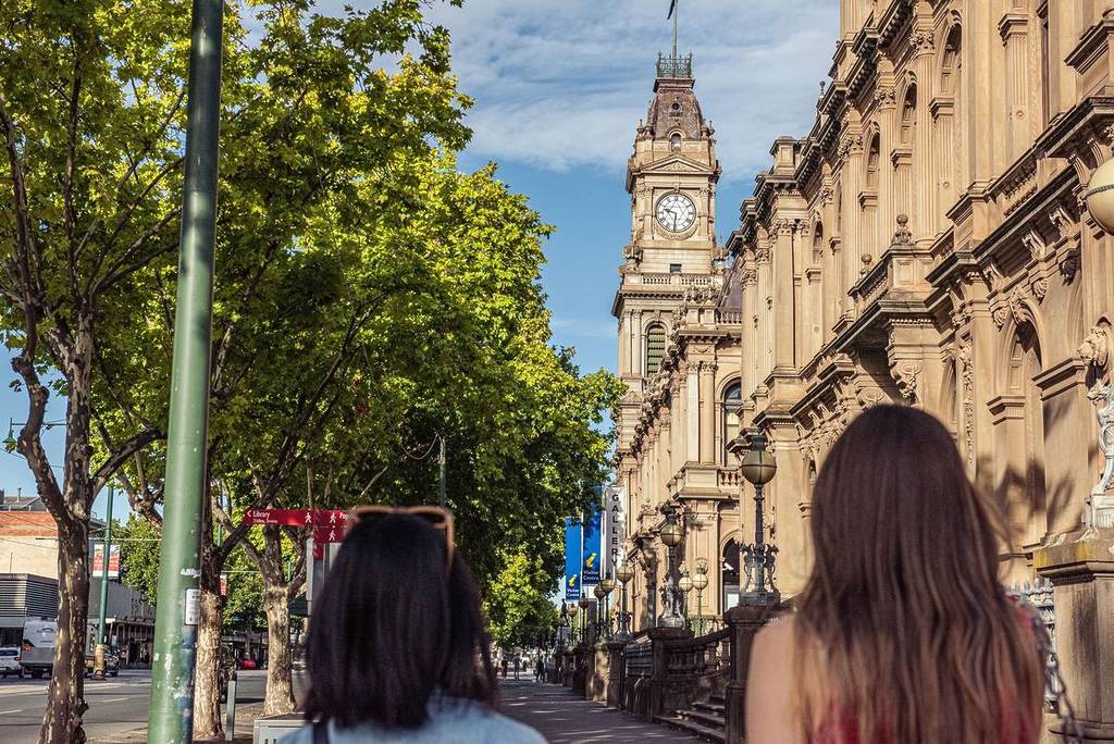 A view of Bendigo's grand clock tower, framed by trees, with two people walking along the historic street