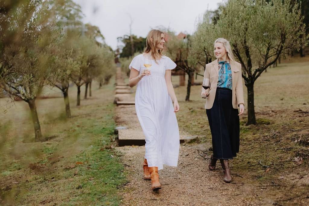 two women holding wine glasses and walking through a vineyard