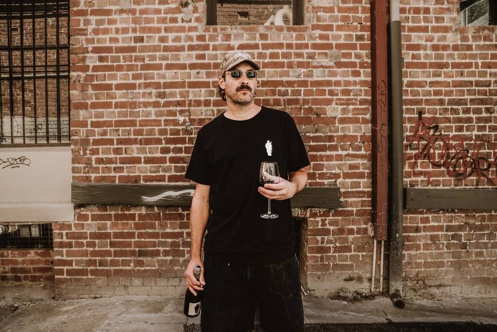 man holding glass of Innocent Bystander wine in front of a graffitied brick wall
