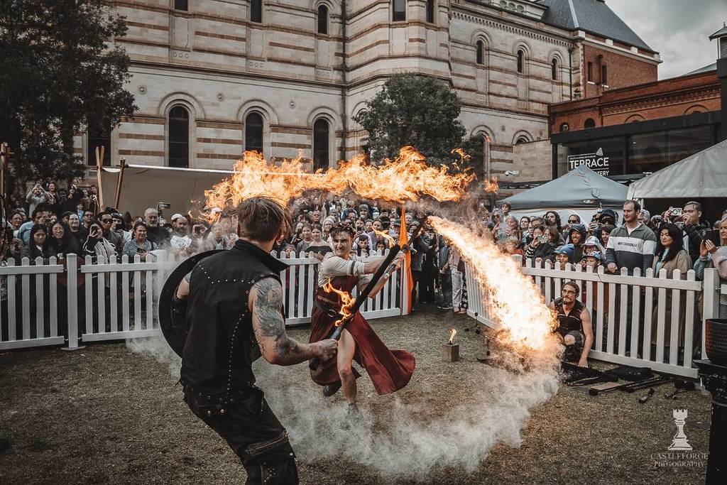 a female fire performer battling a man holding a sword and shield in front of spectators