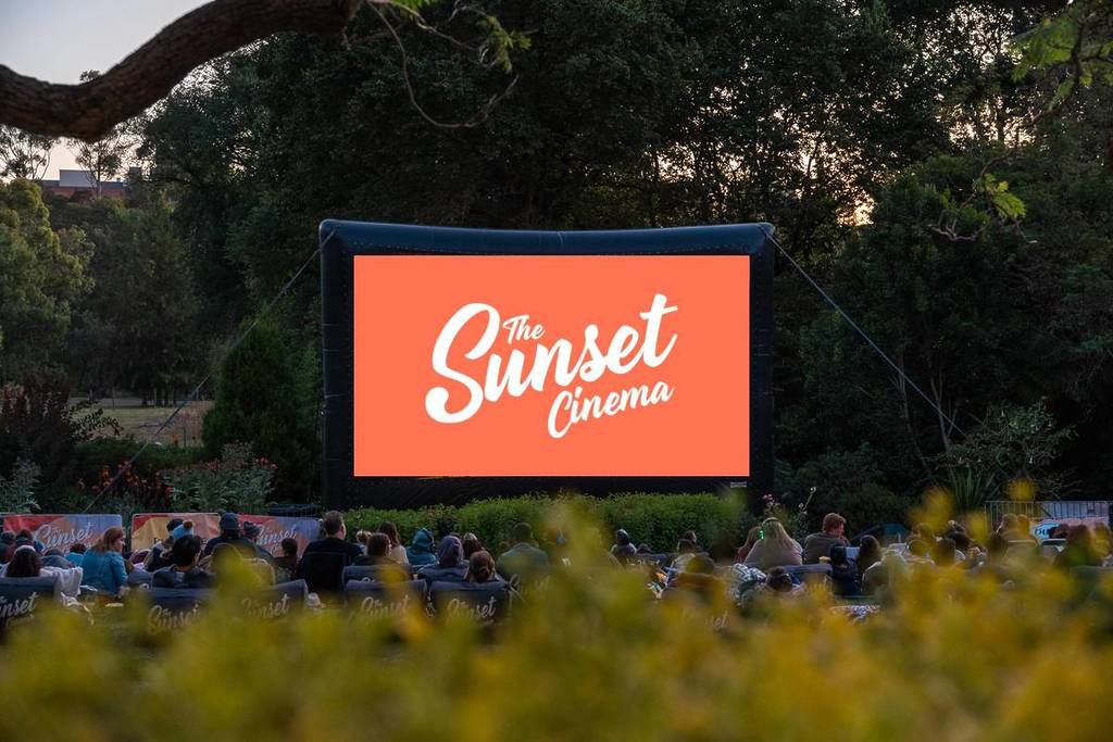 spectators in a park looking at a screen that says Sunset Cinema