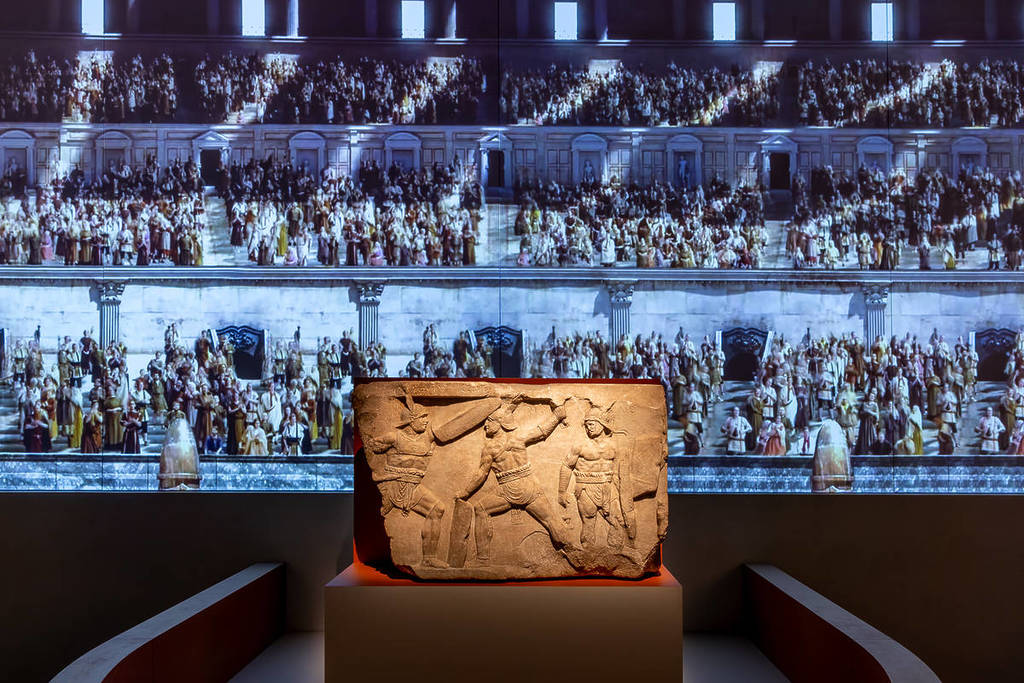 a stone tablet depicting warriors fighting with a projection in the background of a gladiator arena in the Rome exhibition at Melbourne Museum