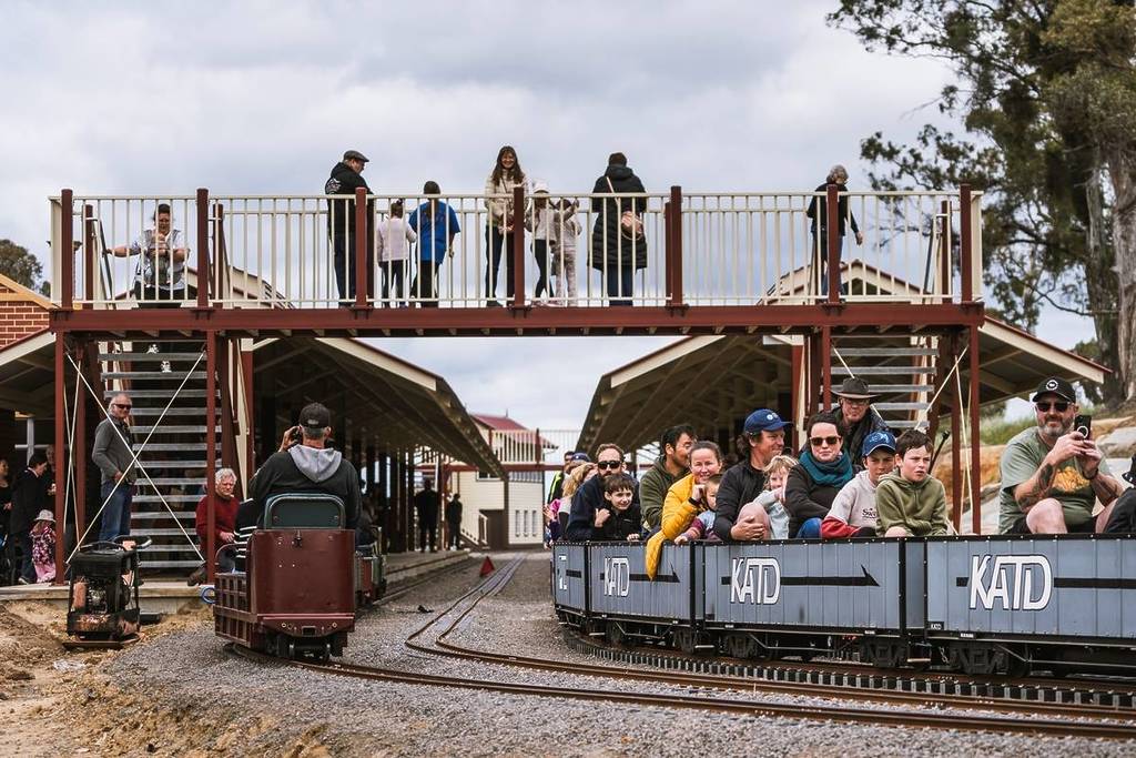 families riding a mini train under a bridge with onlookers at Victorian Miniature Railway