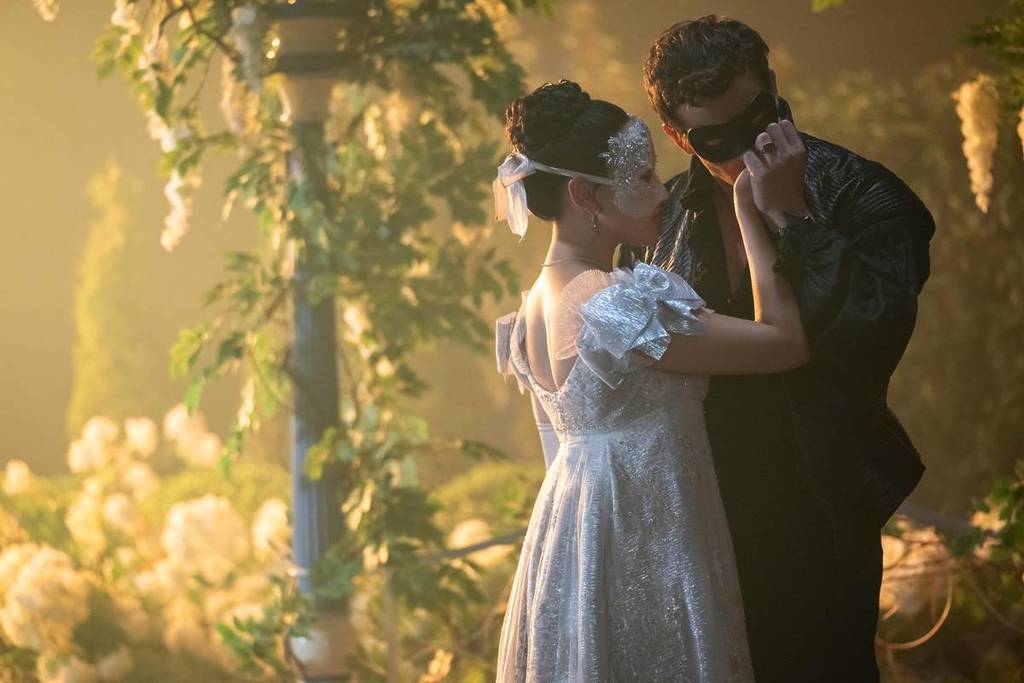 Yerin Ha as Sophie and Luke Thompson as Benedict wearing masks at a romantic ball in Bridgerton, Benedict is kissing Sophie's hand