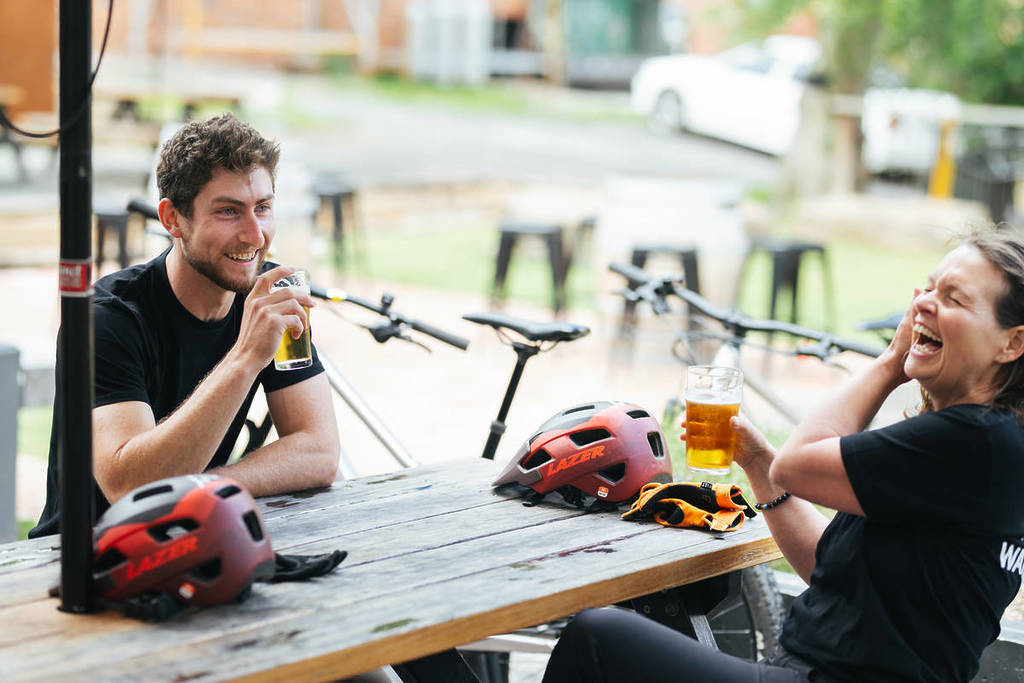 man and woman laughing and sipping beer with bike helmets on a picnic table