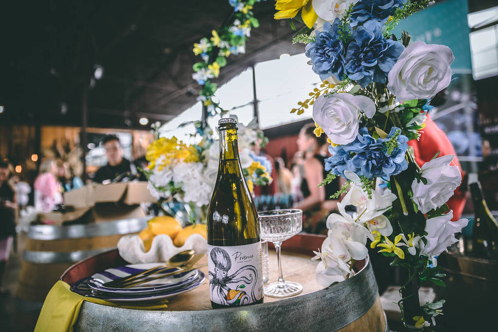 a bottle of wine on a barrel with a floral arch in the background at Wine & Cheese Fest