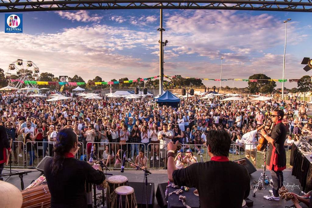 view from on stage behind a band performing at Momo Fest, looking out at crowds, with the tops of food stalls and a Ferris wheel visible in the distance