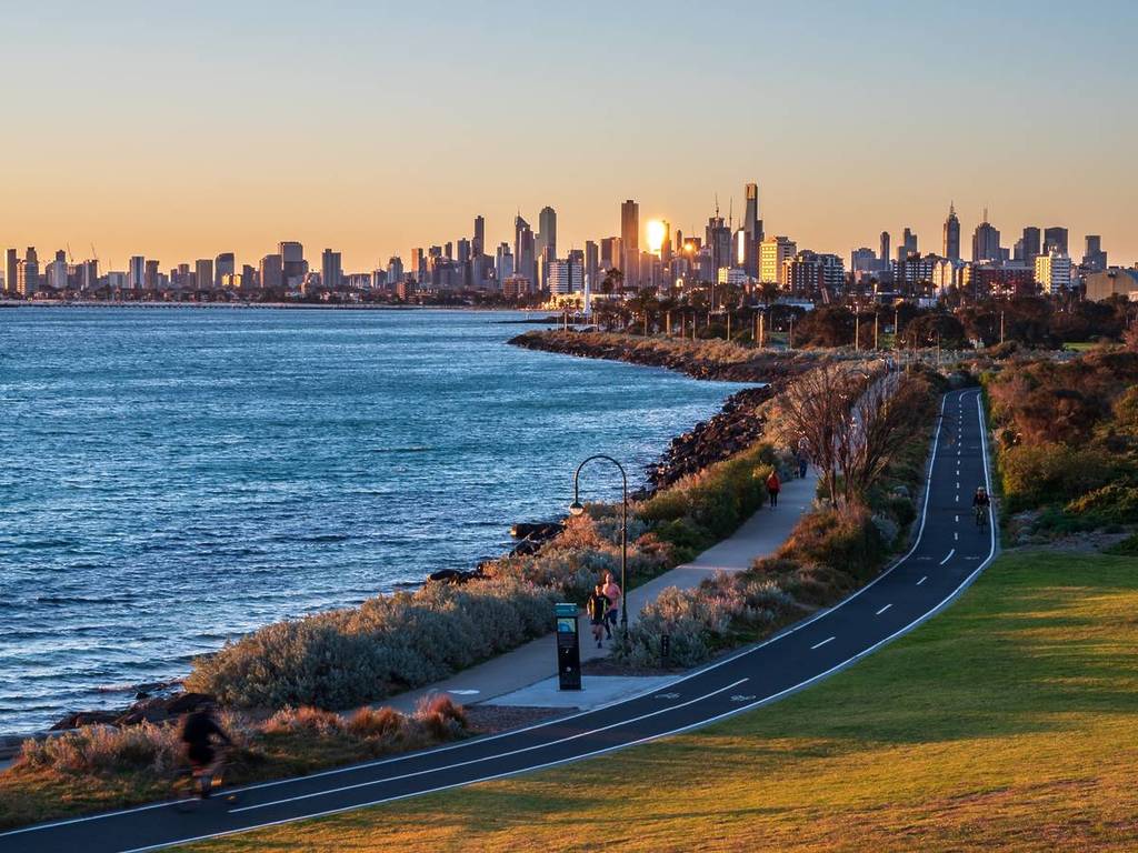 the city skyline at sunset, with light reflecting from one of the buildings, while joggers exercise on a path along the bay, as seen from Point Ormond