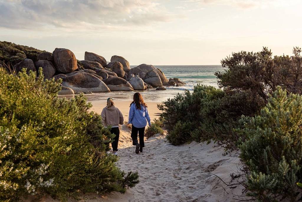 two women walking on the sand to Whisky Bay, with granite boulders in the background and the sea just slightly visible