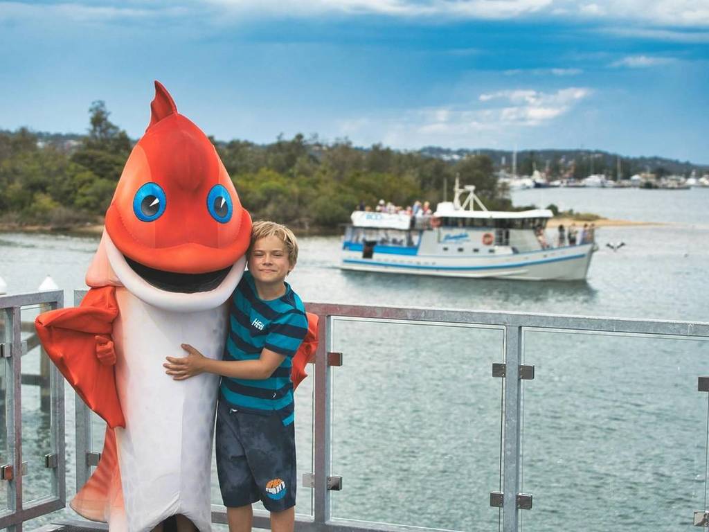boy hugging someone dressed in a fish costume with a boat in the background at Lakes Entrance