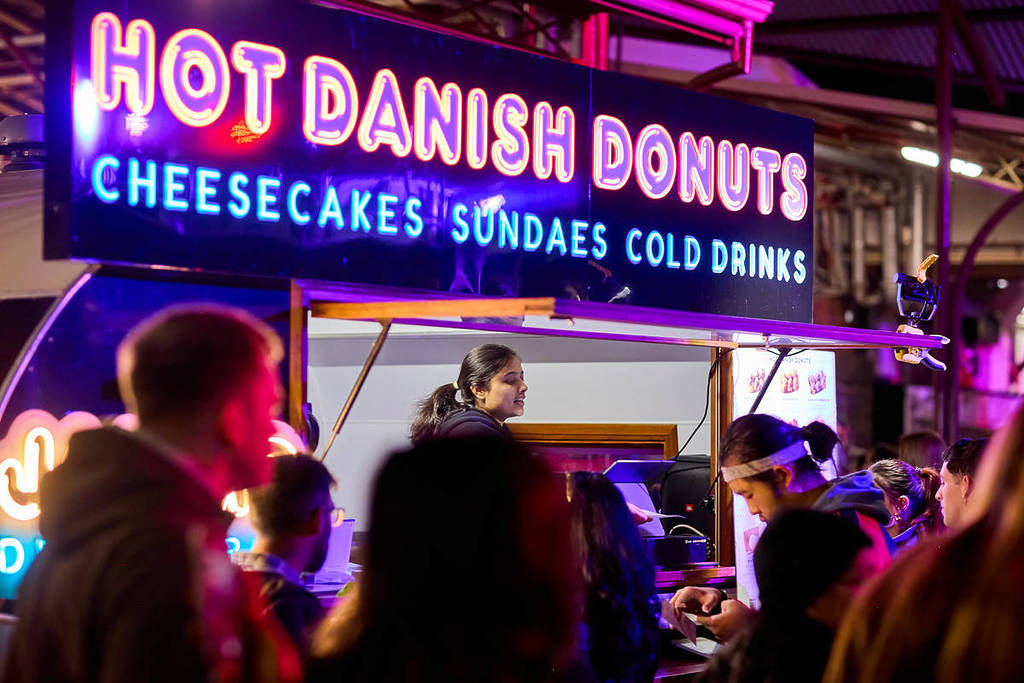 someone waiting to serve a customer in a food truck with a neon sign showing that they sell hot Danish donuts
