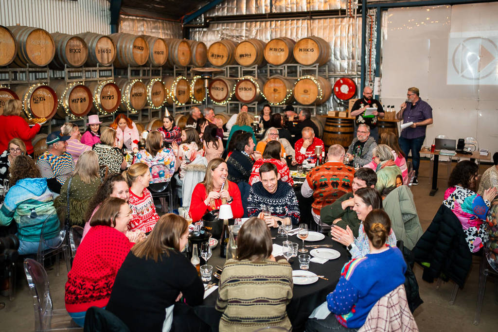 An event inside a cellar at Dark Side of Wine. Groups of people in funny sweaters sit around tables chatting and drinking wine.