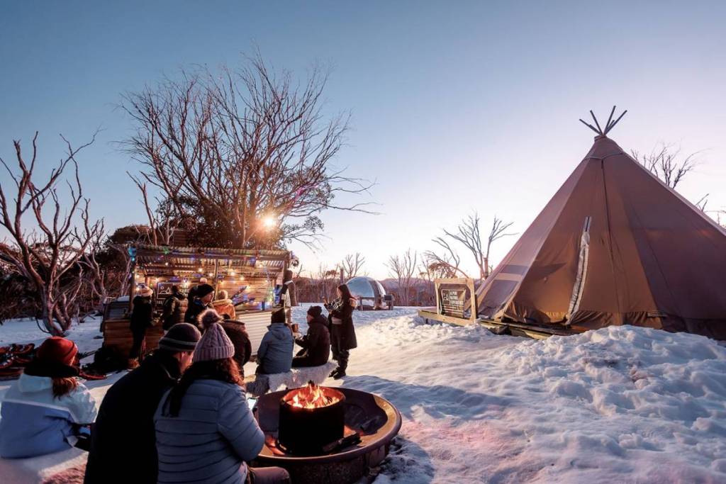 people hanging out by the fireplace, with the tipi and outdoor bar in the background, at the eco-village from Alpine Nature Experience