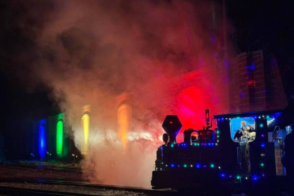 a mini train from Diamond Valley Railway passing a building lit up in colours for Winter Light Spectacular