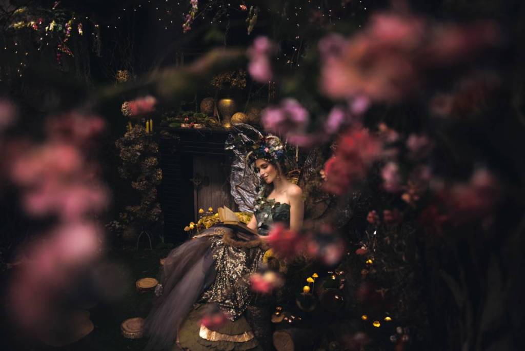 woman dressed as dark fairy reading a book, surrounded by flowers