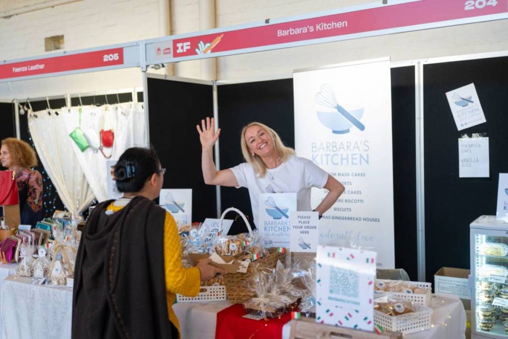 woman at a market stall selling baked goods waving to camera