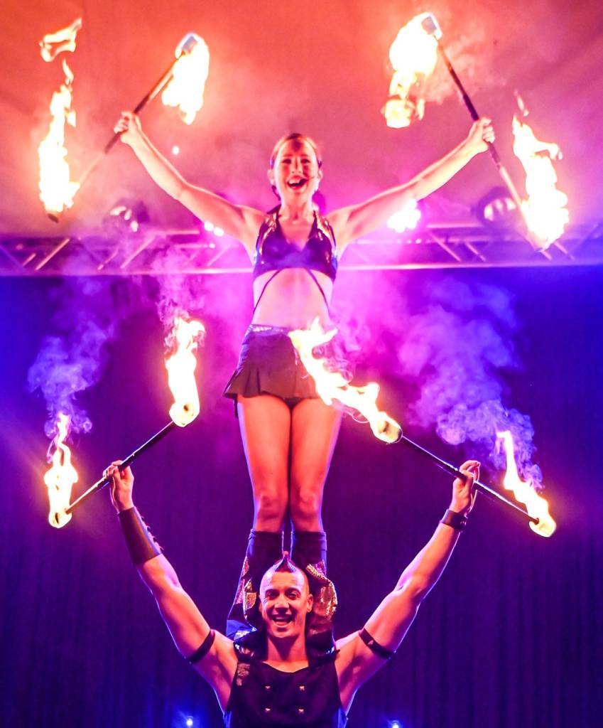 one performer standing on another's shoulders, both holding fire sticks