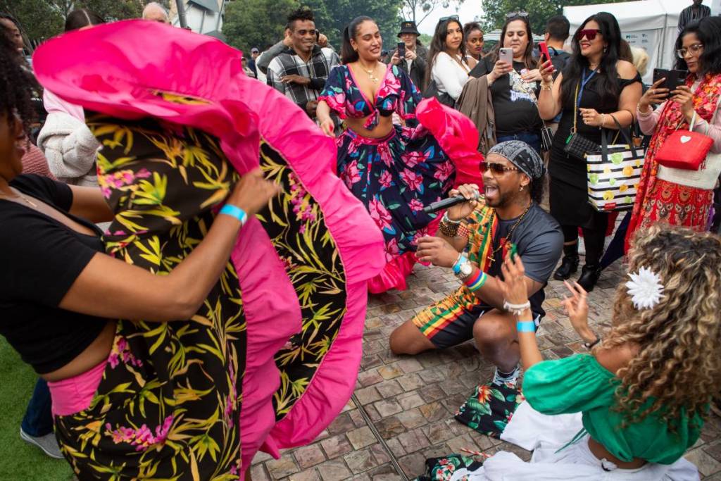 man crouching with a mic while woman dances with large skirt, surrounded by energetic crowd at African Music and Cultural Festival