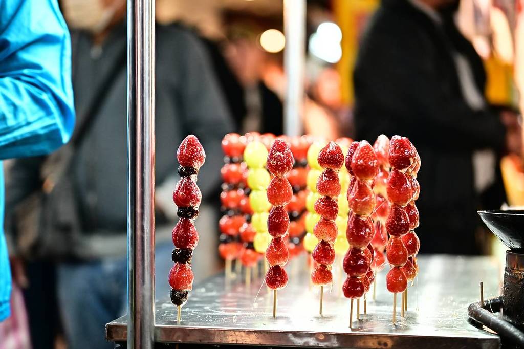 Contra o pano de fundo de um animado mercado noturno de inverno, uma barraca de tanghulu brilha suavemente. As delicadas cascas de açúcar brilham sob as luzes do mercado, criando um ambiente nostálgico e festivo.