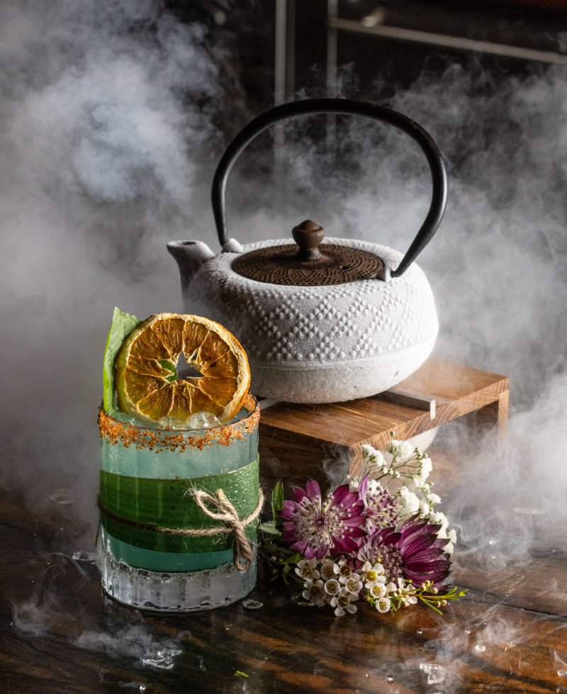 a green-colored cocktail with a Japanese tea kettle in the background, and smoke rising around both