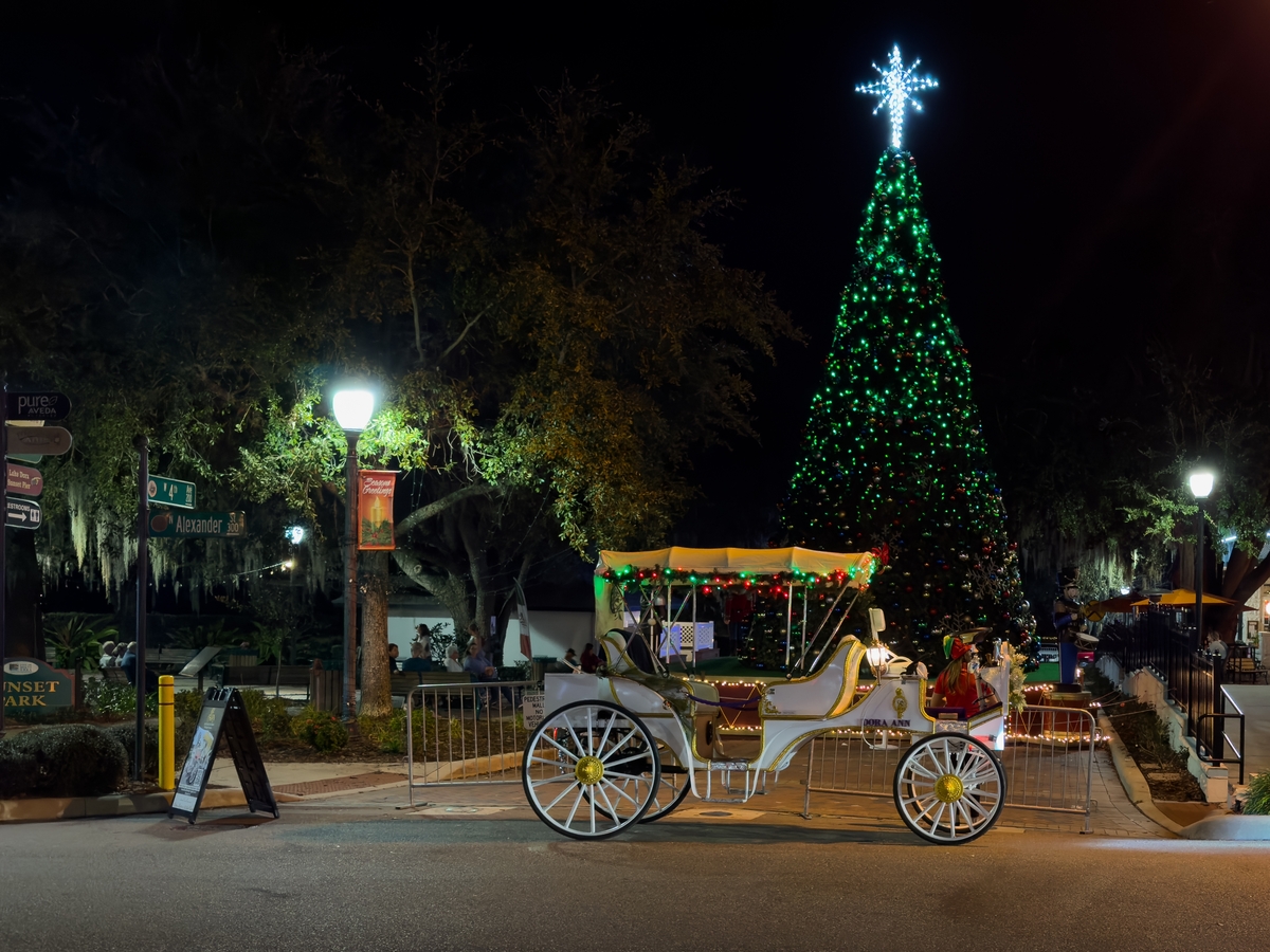 Mount Dora, Florida, US-November 1, 2023: A motorized white carriage parked in a town square with a driver in traditional dress, a tall Christmas tree with green lights, baubles, and a nutcracker doll